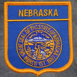 May include: A blue and gold embroidered patch featuring the word "NEBRASKA" at the top. The patch has a shield shape with an orange border. The center features a circular emblem with the words "GREAT SEAL OF THE STATE OF NEBRASKA" and the date "MARCH 1ST 1867".