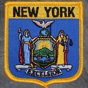 May include: Embroidered patch featuring the New York state seal. The patch has a blue background with a yellow border. The words "NEW YORK" are at the top. The state seal includes an eagle, two figures, and the word "EXCELSIOR".