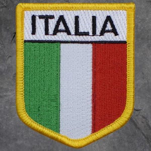 May include: Embroidered patch featuring the Italian flag design. The patch has a shield shape with a yellow border. The Italian flag colors of green, white, and red are vertically striped. The word "ITALIA" is embroidered in black letters at the top.