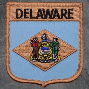 May include: Embroidered patch featuring the word "DELAWARE" in black block letters above a light blue shield. A diamond shape in the center displays the state seal with figures and a coat of arms, all outlined in brown.