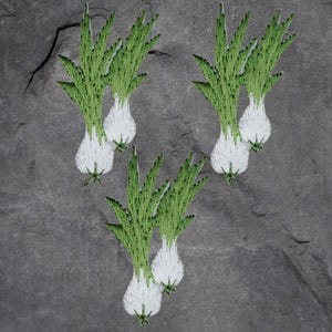 May include: Embroidered patches featuring green and white scallions. The patches are detailed with green leaves and white bulbs, arranged on a gray, textured background. These patches could be used to decorate clothing or accessories.