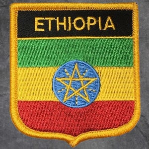 May include: Embroidered patch featuring the Ethiopian flag design. The patch is shield-shaped with a gold border. The flag design includes horizontal stripes of green, yellow, and red, with a blue emblem in the center. The word "ETHIOPIA" is embroidered in gold.
