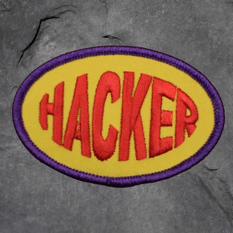 May include: Embroidered patch featuring the word "HACKER" in red, bold letters. The text is on a yellow oval, framed by a purple border. The patch is designed for clothing or accessories.