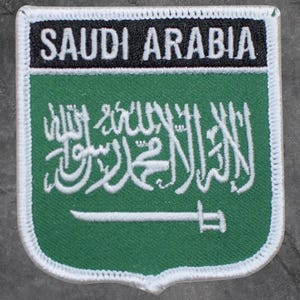 May include: A green and white embroidered patch with the text "SAUDI ARABIA" at the top. The patch features the Saudi Arabian flag design, including Arabic script and a sword.