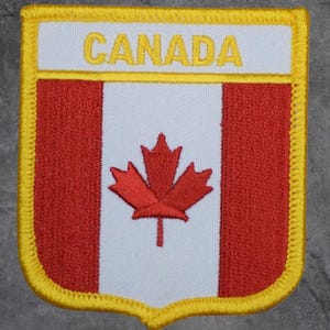 May include: Embroidered patch featuring the Canadian flag design. The patch has a white background with a red maple leaf and two red vertical stripes. The word "CANADA" is embroidered in yellow at the top, and the patch is outlined in yellow.