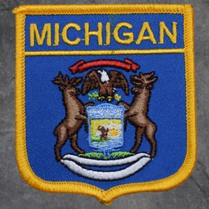 Peut inclure: Écusson brodé bleu et or avec le mot "MICHIGAN" en jaune. L'écusson représente le sceau de l'État avec deux cerfs, un aigle et un bouclier. L'écusson a une bordure jaune.