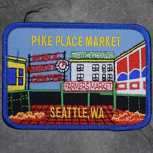 May include: Embroidered patch depicting Pike Place Market in Seattle, WA. The design includes the text "Pike Place Market", "Public Market Center", "Farmers Market", and "Seattle, WA" in various colours against a blue background.