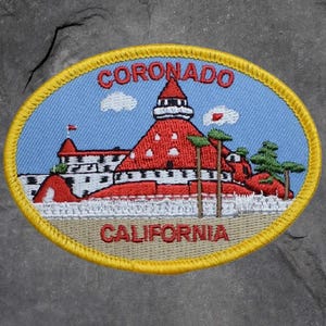 May include: Embroidered patch featuring a red building with a white top and blue sky background. The patch is oval-shaped with the words "CORONADO" and "CALIFORNIA" in red. The patch has a yellow border and a tan base.