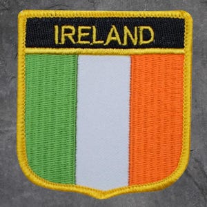 May include: Embroidered patch featuring the flag of Ireland. The patch has a shield shape with a gold border. The flag design includes vertical stripes of green, white, and orange, with the word "IRELAND" in gold letters on a black background.