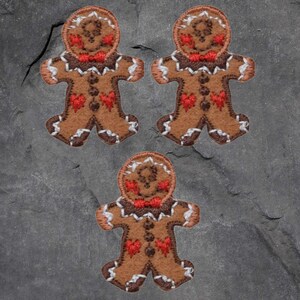 May include: Three gingerbread man patches. Each patch is brown with white trim, red heart accents, and a red bow tie. The patches are arranged on a grey, textured surface.