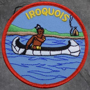 May include: Embroidered patch with a circular design featuring the word "IROQUOIS" in yellow letters. The patch depicts a Native American in a canoe on blue water, with a green shoreline and a small teepee in the background. The patch has a red border.