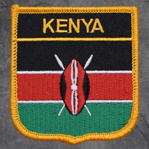 May include: Embroidered patch featuring the word "KENYA" in yellow on a black background. Below, the patch displays the Kenyan flag's colors: black, red, and green, with a shield and spears emblem in the center. The patch has a yellow border.