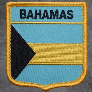 May include: Embroidered patch featuring the flag of the Bahamas. The patch has a light blue background with a yellow horizontal stripe and a black triangle. The word "BAHAMAS" is embroidered in black at the top, with a gold border.