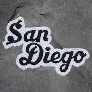 May include: Embroidered patch featuring the words "San Diego" in a black script font with a white border. The patch is placed on a gray, textured surface.