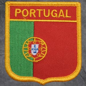 May include: Embroidered patch featuring the flag of Portugal. The patch is shield-shaped with a green vertical stripe on the left and a red vertical stripe on the right. The word "PORTUGAL" is embroidered in yellow at the top.