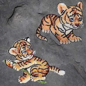 May include: Two embroidered iron-on patches of orange and black tiger cubs. One cub is sitting and holding a green stick. The other cub is standing with its paws outstretched.