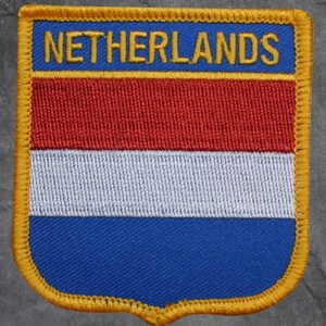 May include: Embroidered patch featuring the Netherlands flag design. The patch has a blue top section with the word "NETHERLANDS" in yellow. Below are horizontal stripes of red, white, and blue, with a gold border.
