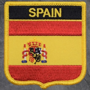May include: Embroidered patch featuring the flag of Spain. The patch has a shield shape with a yellow border. The top section is black with the word "SPAIN" in yellow. The flag design includes red and yellow horizontal stripes, and a coat of arms.