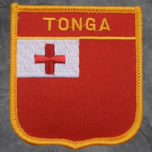 May include: A rectangular patch featuring the flag of Tonga. The patch has a red background with a white square in the upper left corner containing a red cross. The word "TONGA" is embroidered in yellow at the top.