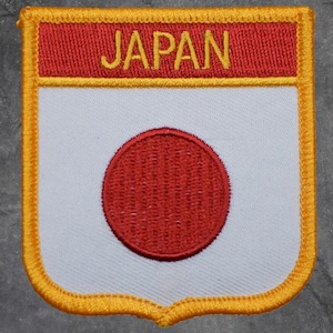 May include: Embroidered patch featuring the flag of Japan. The patch has a white background with a red circle in the center, and a red rectangle at the top with the word "JAPAN" in yellow. The patch is bordered with gold stitching.