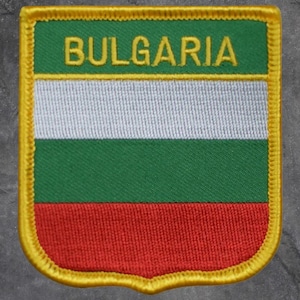 May include: Embroidered patch featuring the flag of Bulgaria. The patch has a shield shape with a yellow border. The flag design includes horizontal stripes of white, green, and red, with the word "BULGARIA" embroidered in gold.