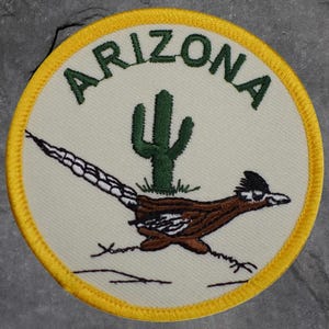 May include: Embroidered patch featuring the word "ARIZONA" in green above a green cactus and a brown roadrunner. The patch has a cream-colored background with a yellow border.