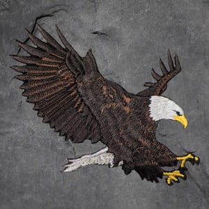 May include: Embroidered patch featuring a bald eagle in flight. The eagle has brown and tan feathers, a white head, and a yellow beak and talons. The patch is set against a dark grey background.