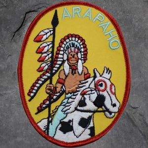 May include: Embroidered patch with a yellow background and a red border. The patch features an illustration of a Native American chief on horseback, holding a spear. The word "ARAPAHO" is arched across the top in light blue.