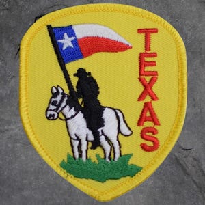 May include: A yellow embroidered patch featuring a rider on a white horse holding a Texas flag. The flag is blue with a white star and red and white stripes. The word "TEXAS" is embroidered in red.