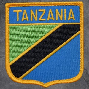 May include: Embroidered patch featuring the word "TANZANIA" in yellow on a blue background. The patch has a shield shape with a green and black diagonal stripe, outlined in yellow and a gold border.