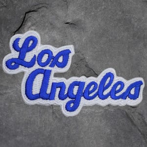 May include: A blue and white embroidered patch with the words "Los Angeles" in a cursive font. The text is in blue with a white border, set against a gray stone background.