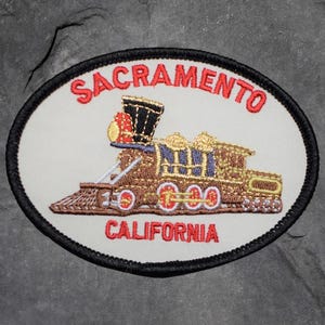 May include: Embroidered patch depicting a detailed vintage train. The patch has a white oval background with the words "SACRAMENTO" and "CALIFORNIA" in red. The train is brown, gold, and blue, with a black border.