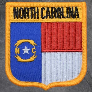 May include: Embroidered patch with the North Carolina state flag design. The patch has a yellow border and the words "NORTH CAROLINA" in black. The flag design incorporates blue, red, and white colours, with a star and the letters "N" and "C".