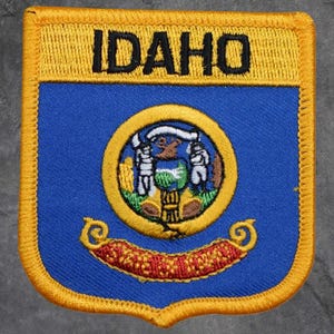 May include: Embroidered patch of the Idaho state seal. The patch has a blue background with a gold border and the word "IDAHO" in black. The state seal is in the centre, with a gold ring and a red and gold banner at the bottom.