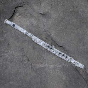 May include: A crocheted flute in shades of gray and white, with dark gray accents on the keys. The instrument is lying diagonally on a dark gray, textured surface. The flute's design includes detailed crochet work.