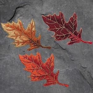 May include: Three embroidered autumn leaves in shades of orange, red, and brown on a grey background.