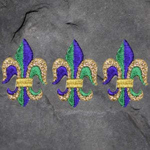 May include: Three embroidered fleur-de-lis patches. Each patch features a gold glitter outline, with sections of green, purple, and gold. The patches are arranged in a row against a dark gray, textured background.