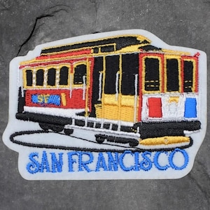 May include: Embroidered patch featuring a San Francisco cable car. The patch has a white border and includes the words "SAN FRANCISCO" in blue. The cable car is red, yellow, black, and white.