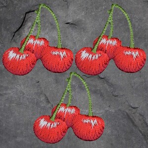 May include: Embroidered patches of red cherries with white detailing and green stems. The patches are set against a grey, textured backdrop. These patches could be used to embellish clothing or accessories.