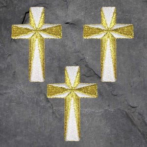 May include: Three embroidered crosses with a gold and white design. The crosses are arranged on a dark gray, textured surface. The gold thread creates a radiant effect, while the white adds contrast.