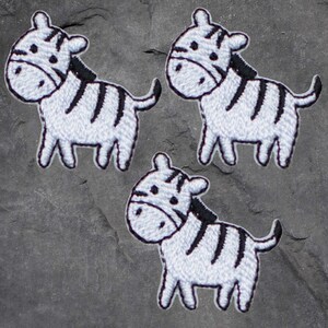 May include: Three embroidered zebra patches. Each patch features a white zebra with black stripes, a black mane, and a small black tail. The patches are on a grey, textured surface.