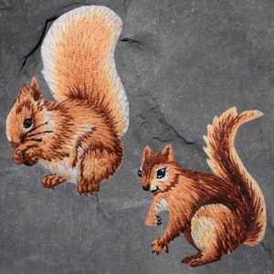 May include: Two embroidered brown and white squirrel patches. The squirrels are facing different directions. The patch on the left has a bushy tail. The patch on the right has a long, thin tail.