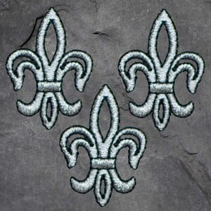 May include: Three embroidered fleur-de-lis patches. Each patch features a silver design with a dark outline, set against a dark gray background. The fleur-de-lis is a stylized lily, a symbol of French royalty.