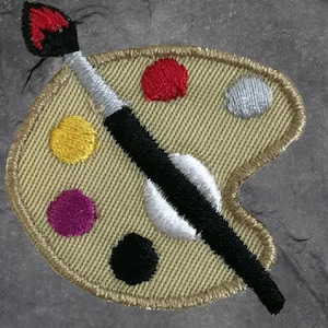 May include: Embroidered patch featuring an artist's palette with a paintbrush. The palette is beige with colorful paint splotches in red, yellow, purple, black, and silver. The paintbrush has a black handle and a silver ferrule.