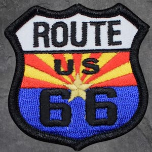 May include: Embroidered Route 66 patch in a shield shape. The top section features the word "ROUTE" in white, above "US" in orange. The middle section has a red and yellow sunburst design. The bottom section has the number "66" in black on a blue background.