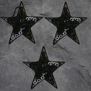 May include: Three black star-shaped patches with green sequins and white accents. The patches are arranged on a grey, textured surface. Each star has a delicate, lacy outline, adding a touch of elegance to the design.