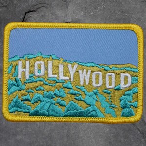 May include: Embroidered patch featuring the word "HOLLYWOOD" in white letters against a backdrop of a blue sky and a yellow-green landscape. The patch has a yellow border and turquoise accents.