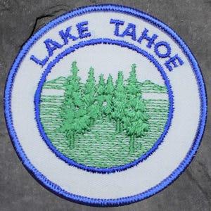 Könnte beinhalten: Gestickter Aufnäher mit den Worten "LAKE TAHOE" in Blau, umgeben von einem grünen und weißen Bild von Kiefern und Bergen. Der Aufnäher hat einen weißen Hintergrund mit blauem Rand.