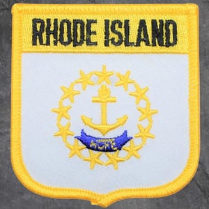 May include: Embroidered patch featuring the text "RHODE ISLAND" in black on a yellow background. The patch has a white center with a gold anchor, blue ribbon, and a ring of gold stars. The patch has a yellow border.