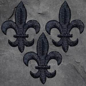 May include: Three black fleur-de-lis patches embroidered on a grey stone surface. Each patch features a detailed, textured design. The fleur-de-lis is a stylized lily, a symbol often associated with royalty and heraldry.